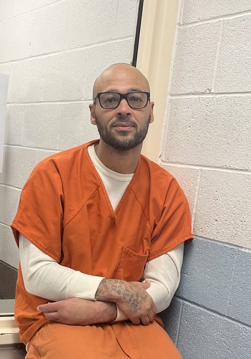 Photograph of a man with glasses in an orange jumpsuit, sitting with his arms crossed.